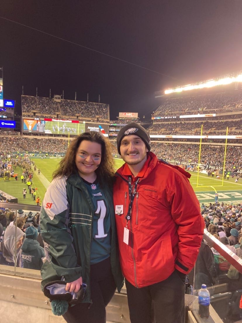 Danny stands with a friend bundled up for an Eagles game as a fan, with the filled stadium behind him. 