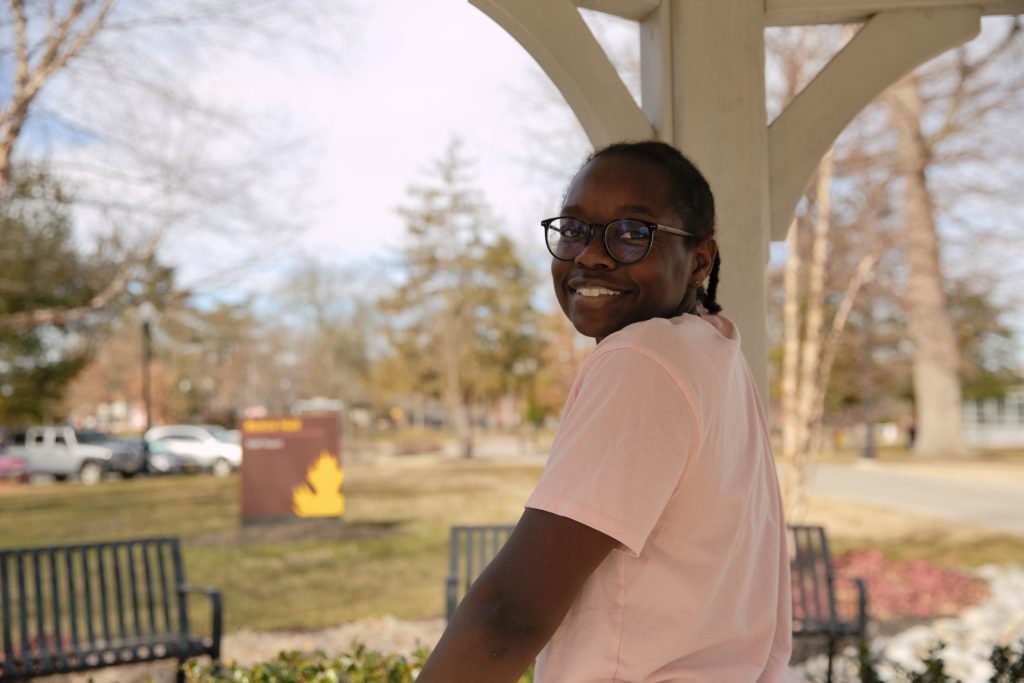 Lila Dasi is posing inside of a pavilion outside of Bunce Hall at Rowan University. Lila is looking over her shoulder, wearing a pink t-shirt, and smiling at the camera. 