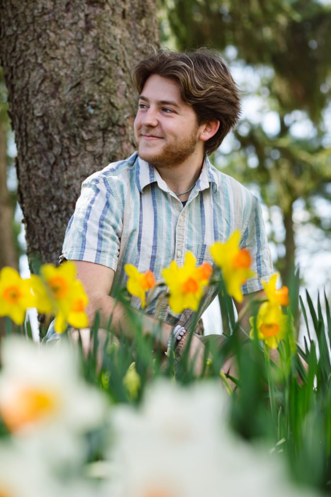 Mark sits in front of a tree with a field of daffodils in front of him. 