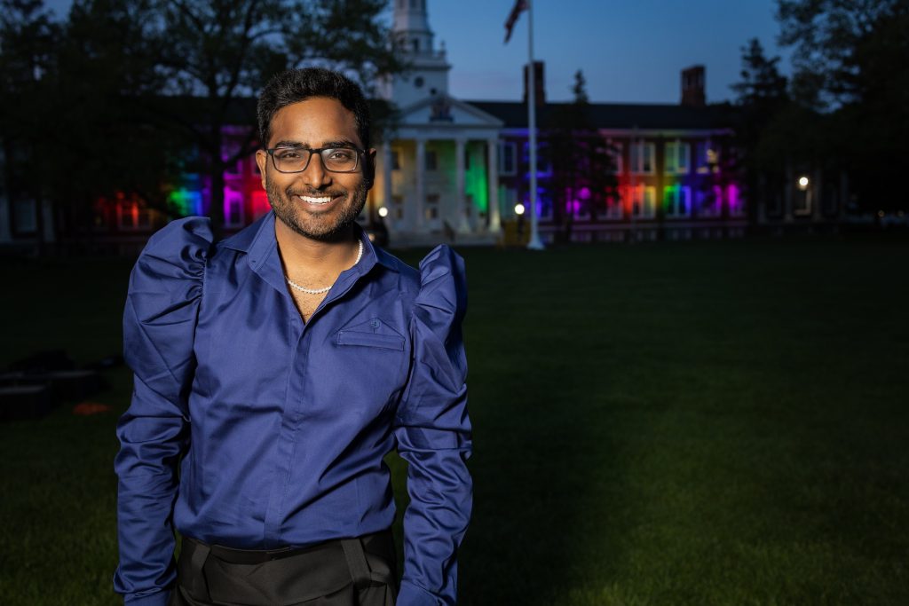 Yash smiling in front of Bunce Hall, which is lit up for Pride month.