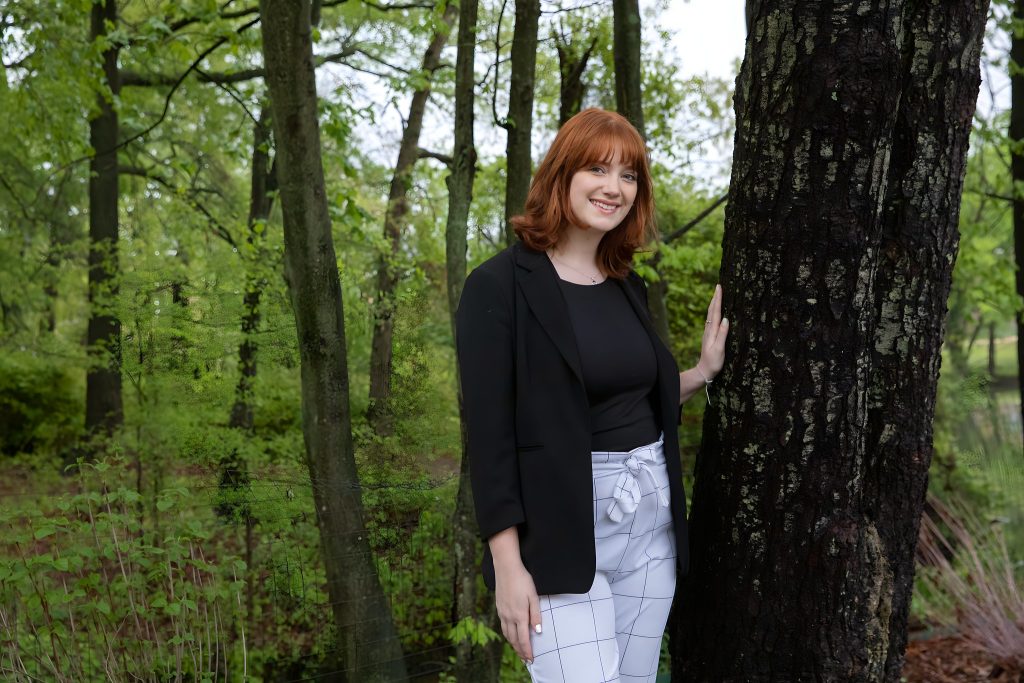 Madelyn smiles while touching a tree, with a wooded background behind her. 