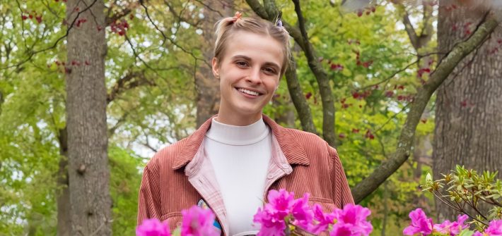 Kit poses for a portrait outdoors, while wearing a bright pink jacket.