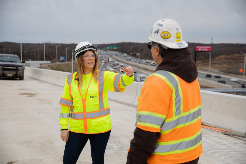 Kate outdoors at a construction site, giving directions to another construction worker, both wearing goggles, bright vests, helmets, and other safety equipment. 