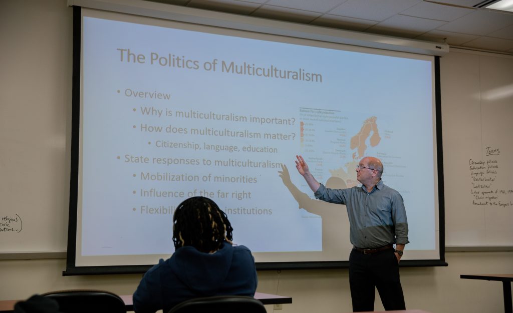 Professor Markowitz stands in front of a slide labed "The Politics of Multiculturalism" with his hand pointing to some of the bullet points such as "Why is multiculturalism important?" and "How does multiculturalism matter?". There is a student in the foreground taking notes.