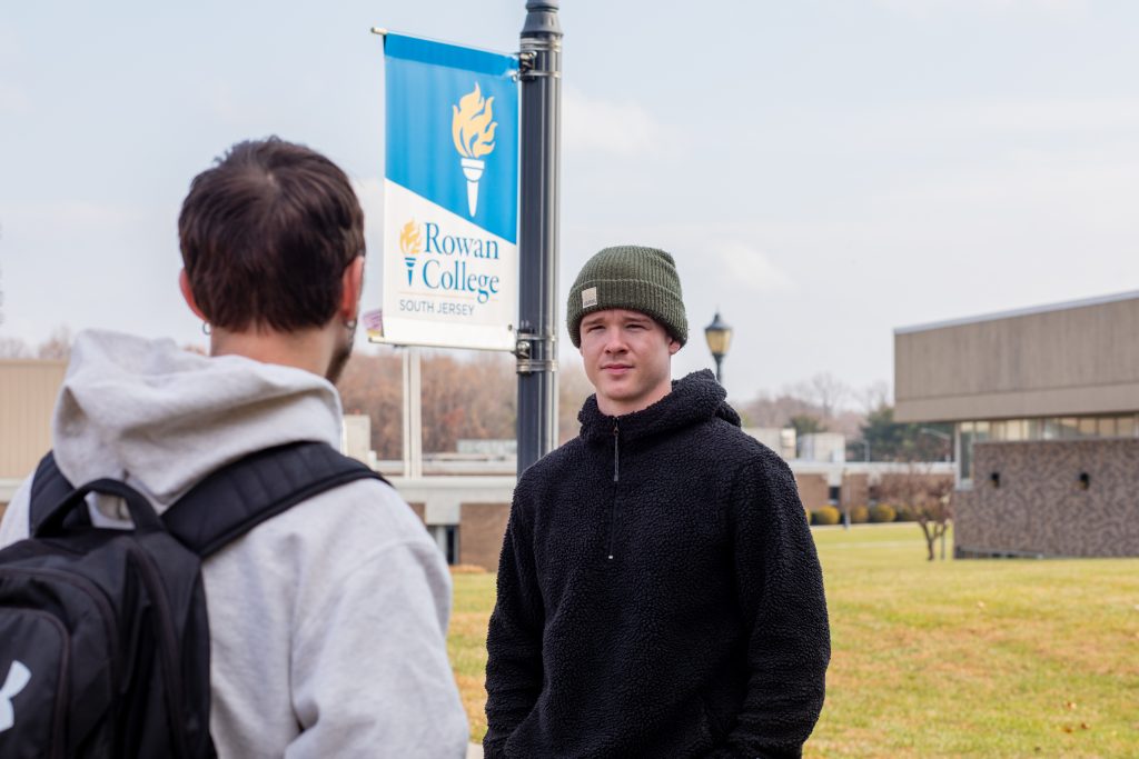 Two students talking in front of a Rowan College of South Jersey flag.
