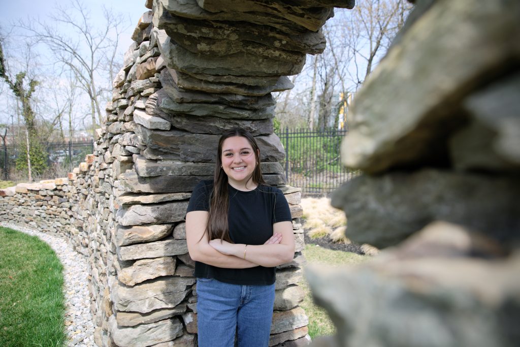 Jean stands under a rock art sculpture on campus, smiling. 