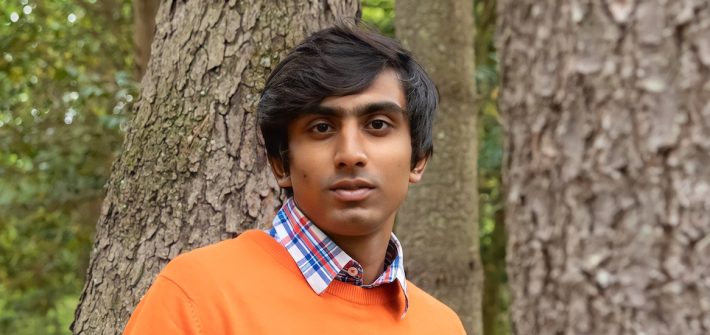 Kriish poses for an outdoor portrait in front of a tree while wearing a bright orange shirt.