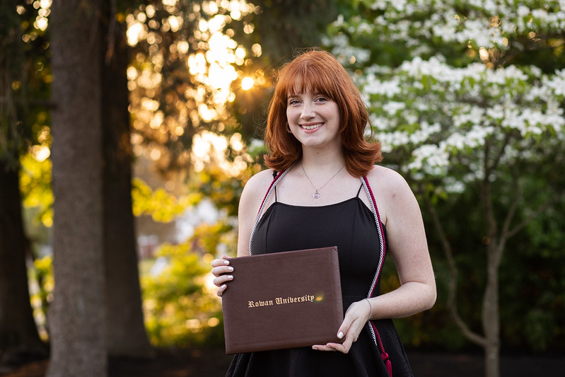 Madelyn standing in front of a sunset with her Rowan diploma.
