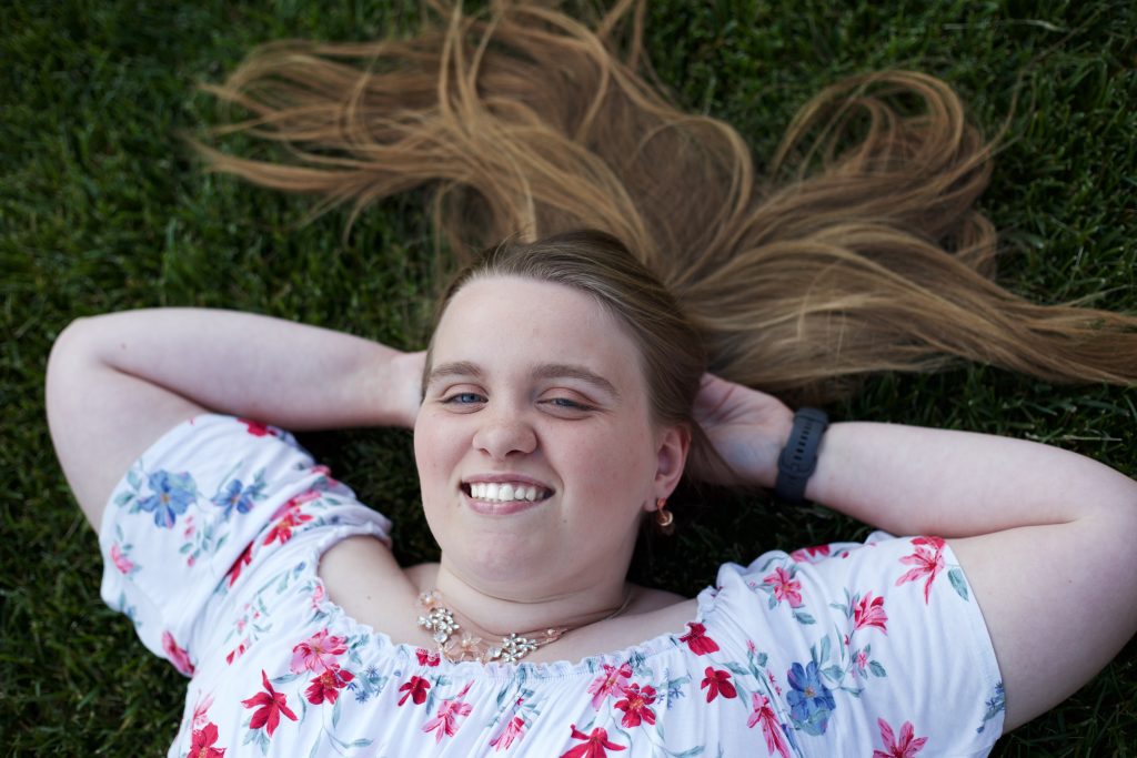Heather is laying down in grass. She is smiling with joy, and she is wearing a white flower dress