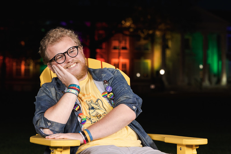 Raymond smiles big relaxing in a yellow chair at night with Bunce Hall behind him. 