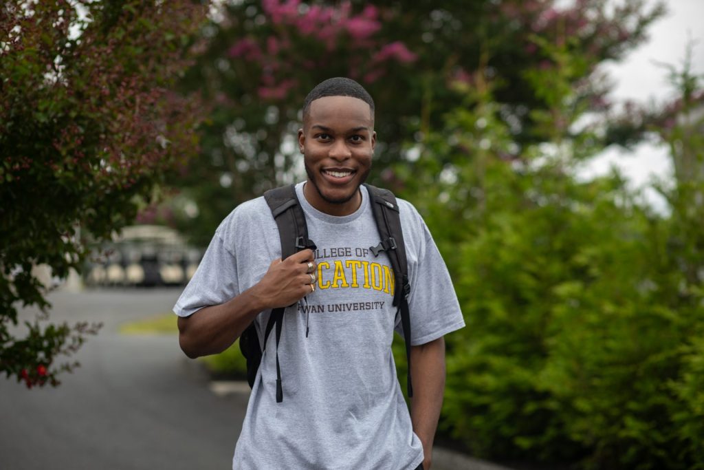 Adam stands on campus with trees behind him, holding onto his backpack with his right hand.