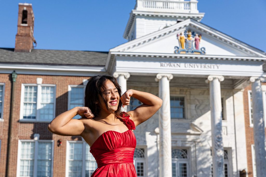 Sapjah Zapotitla stands in a red dress flexing her biceps with Bunce Hall at Rowan University behind her. 