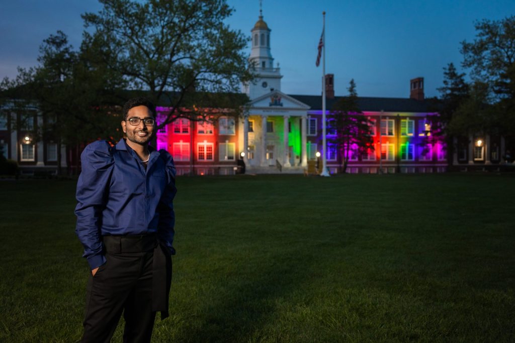 A student stands with hands in his pockets with Bunce behind him illuminated with rainbow lights for Pride. 