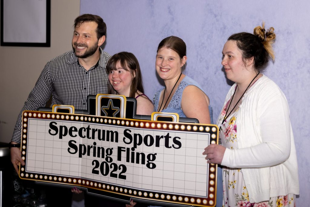 Two athletes stand with two Spectrum Sports staff behind a Spring Fling 2022 sign. 