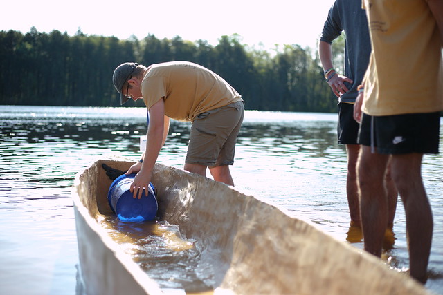 A student uses a blue bucket to scoop water out of the front of the canoe. 