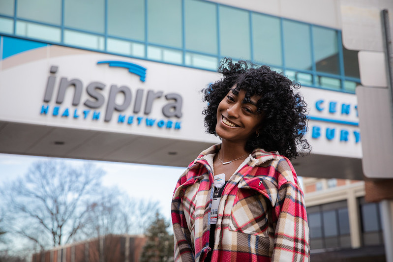 A girl happily tilts her chin while smiling in front of an Inspira sign where she has her internship. 