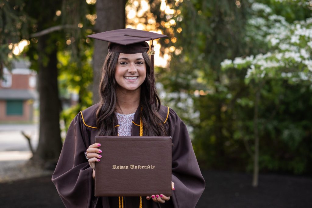 Natalie poses in her brown graduation gown holding her Rowan University diploma holder. 