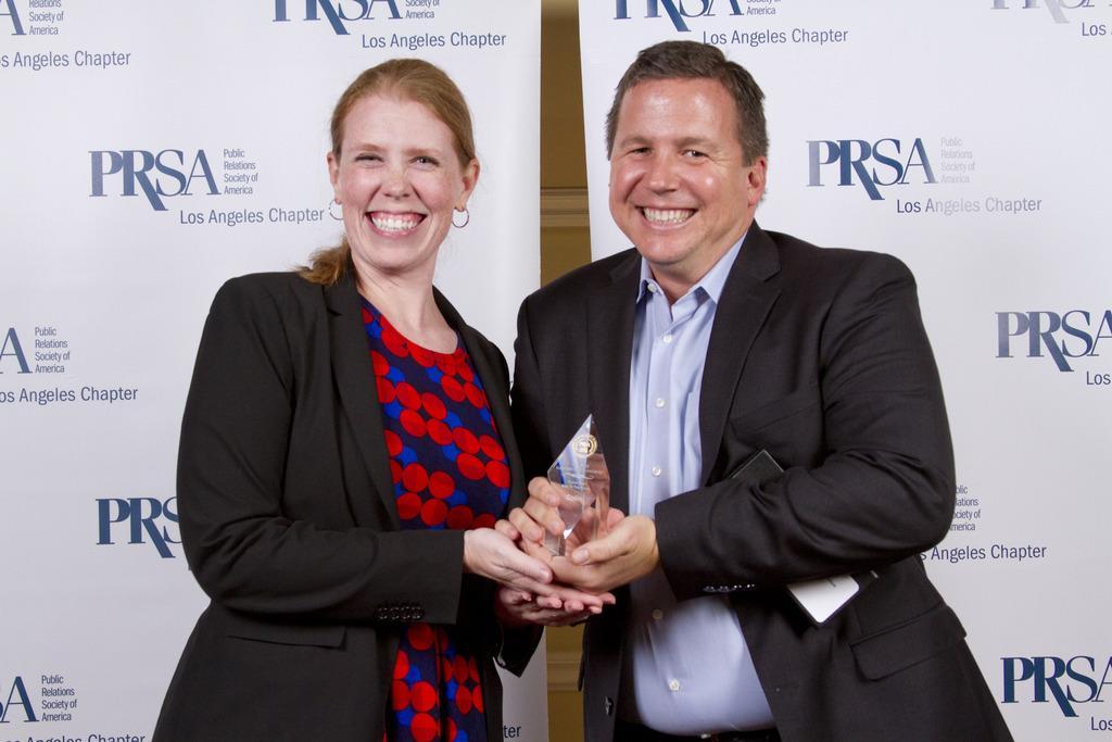 Amy Taylor stands in front of a PRSA professional backdrop at an awards ceremony, presenting a colleague with their award. 