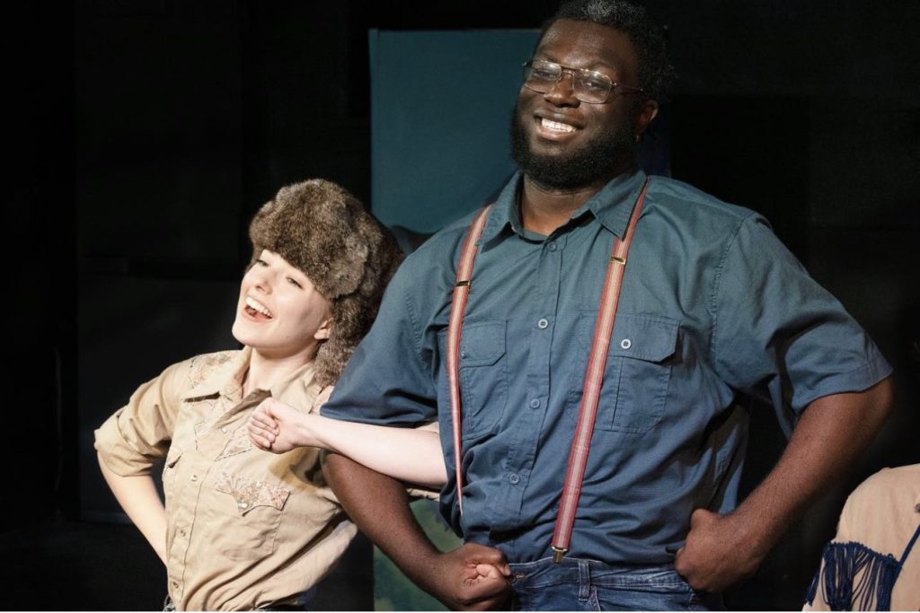 Erica smiles on stage, linked arm in arm with a fellow theatre major during a production.