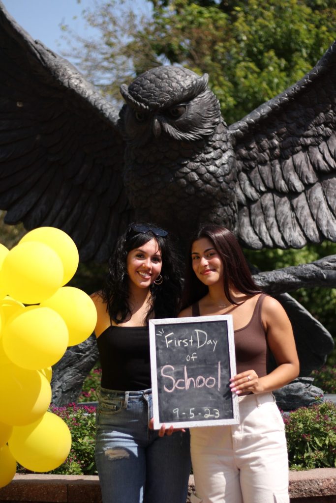 Julianna stands with a friend in front of the iconic owl statue on campus, with yellow balloons by her side and a classic "first day of school" blackboard with chalkboard for the date September 5, 2023. 