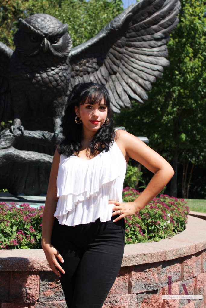 Julianna poses for a beautiful portrait in front of the owl statue on campus, with her hair curled, wearing a white shirt and blank pants. 