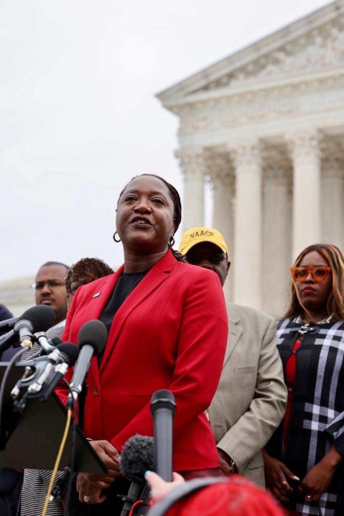 Troi Barnes stands at a podium in front of a bank of media microphones at a Washington, DC, event, while wearing a red suit and talking to the media.