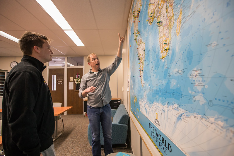 A professor points at a map on the wall, explaining a point to a student. 
