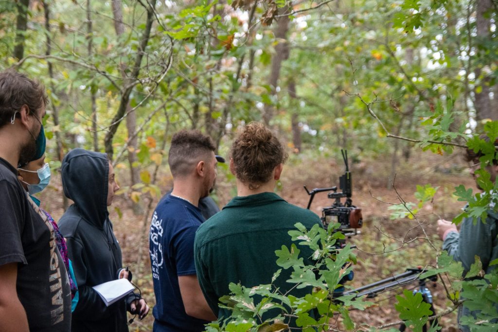 Adam and three crew members examine a camera in the woods while filming. 