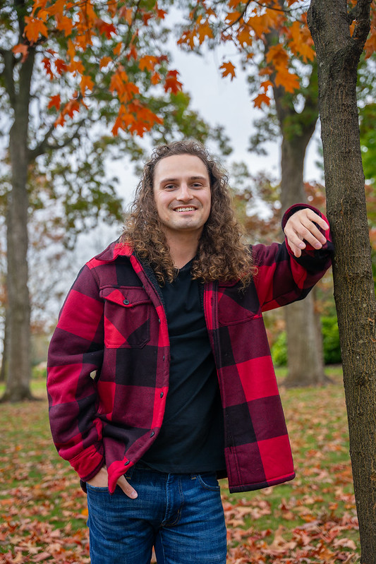 Adam Clark poses outdoors for a portrait, wearing flannel with his arm resting on a tree. 