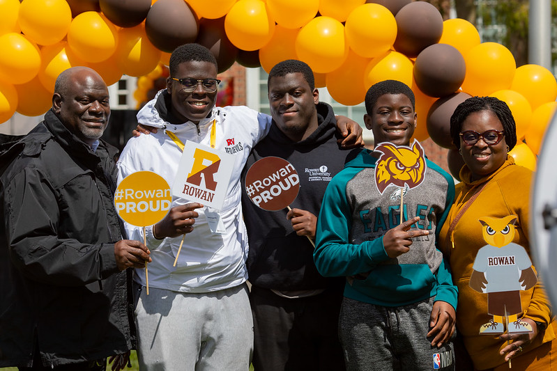 A happy family of five hold #RowanPROUD signs under a brown and gold school color balloon arch at Accepted Students Day. 
