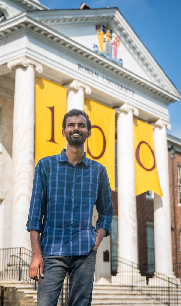 Sai stands in front of the Rowan 100 banner hanging on Bunce Hall. 