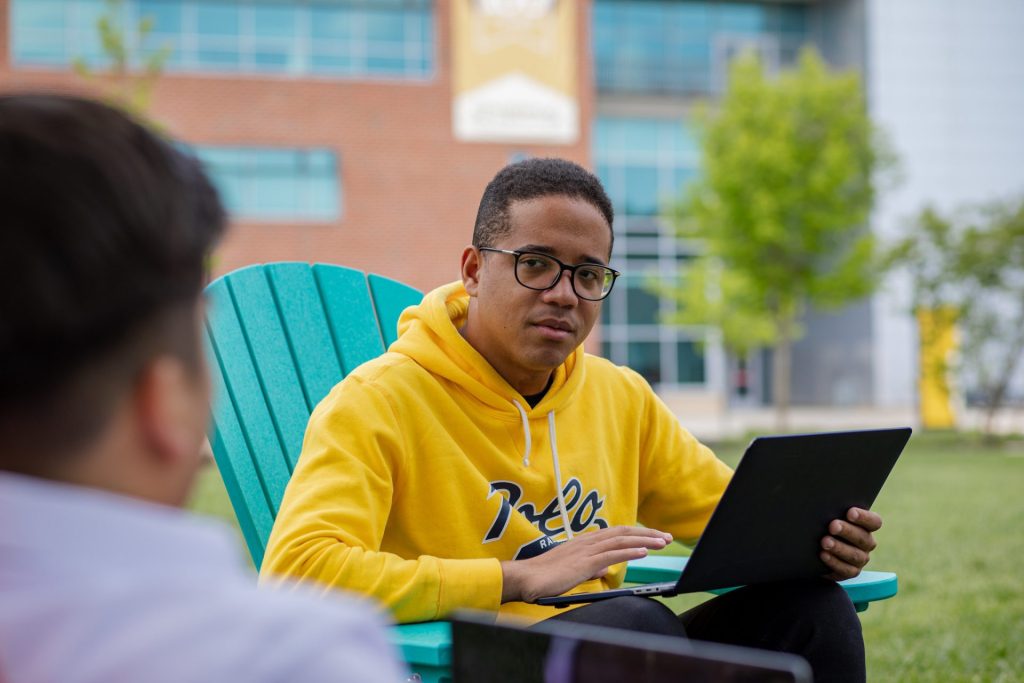 Osvaldo sits outdoors on campus in an Adirondack chair, with a laptop in front of him, talking to a classmate. 