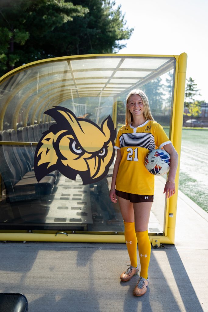 Emily stands casually while resting a soccer ball on her hip, next to an oversized logo of the Rowan mascot, The Prof Whoo RU. 