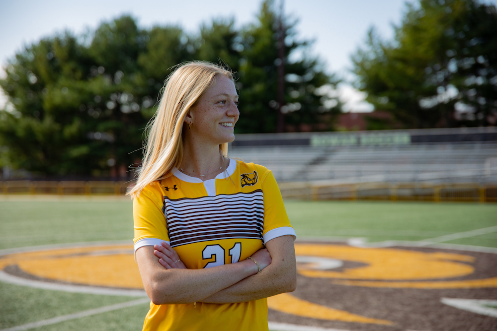 Emily looks off to the side, arms crossed across her chest, while wearing a soccer uniform on the soccer field. 