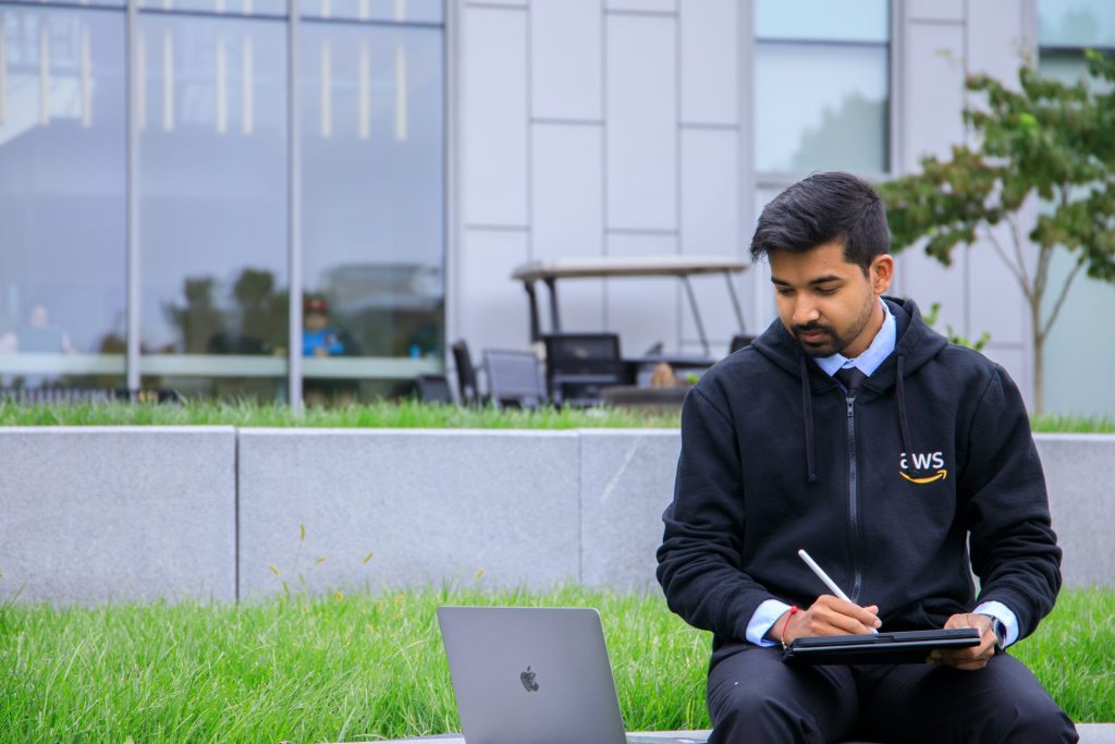 Somyaranjan Rout sits behind business hall, writing in a notebook while looking down at his laptop. 