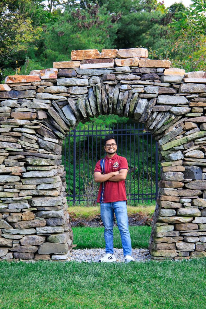 Pintu stands smiling, with his arms across his chest, with a rock sculpture arch behind him. 