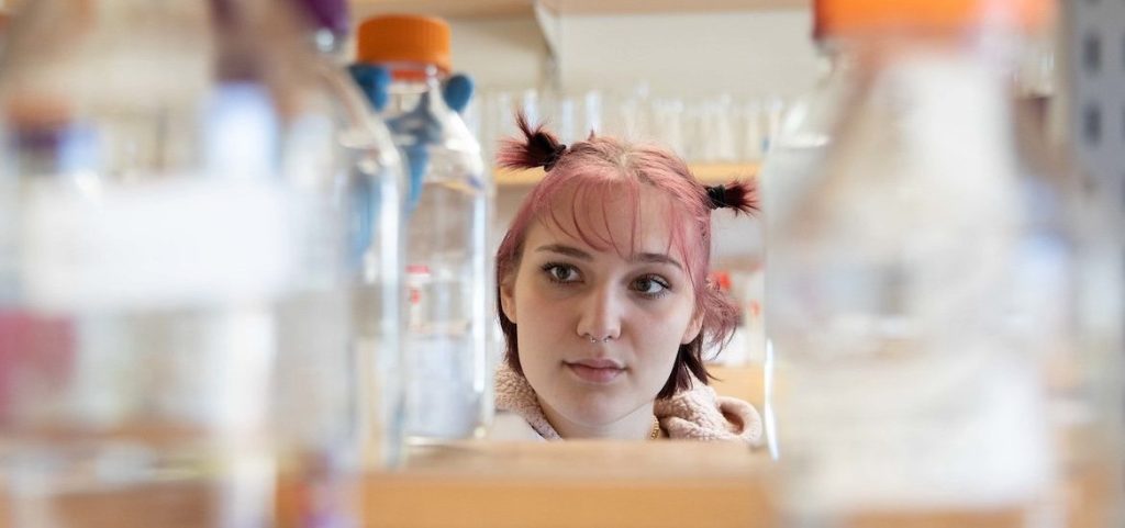 Abigail rocks short pink ponytails, looking through a row of glass bottles on a shelf in a lab. 