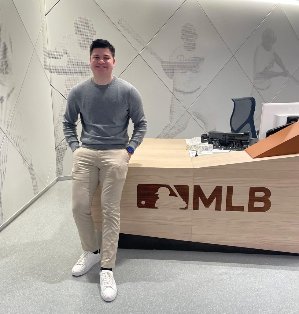 Coby leans casually on an MLB desk at his new job. 
