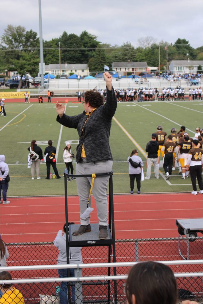 Megan Cooney stands above the field leading the band while looking over her shoulder back to them. 