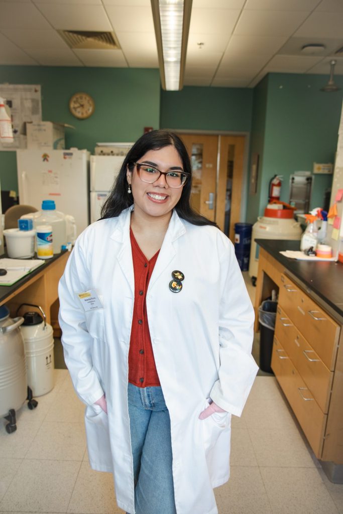 Yesenia stands in a biological sciences lab wearing a white lab coat. 