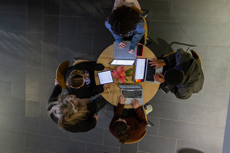An above shot of four students working at a small table with a professor assisting them. 