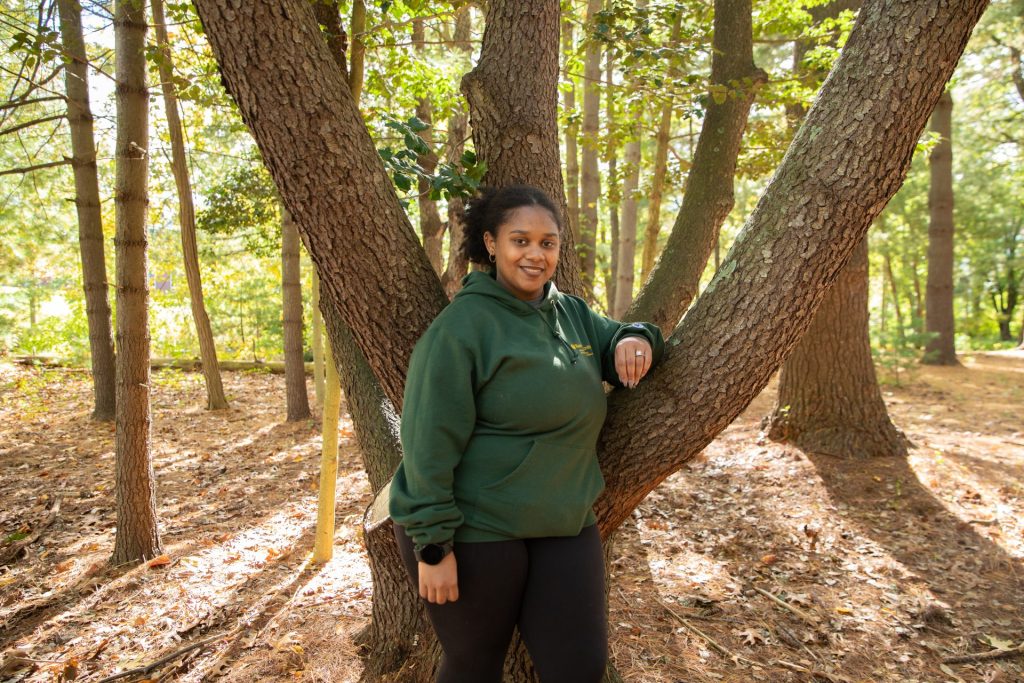 Monica Torres posing outside, leaning on a tree branch. She is wearing a dark green hoodie with her hair pulled back.