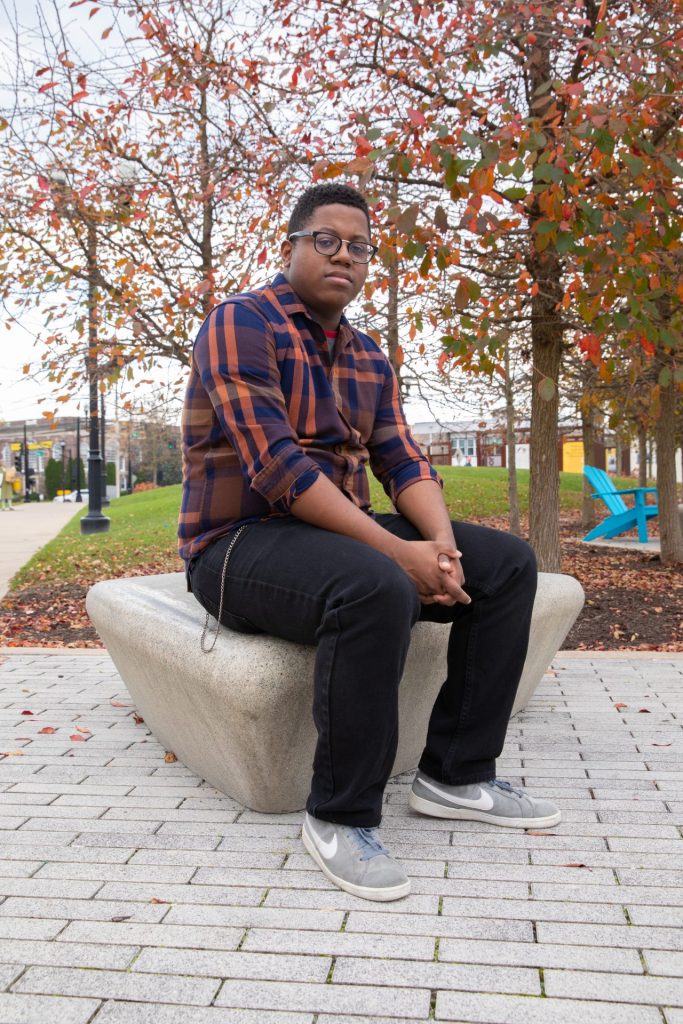 Sedrick sitting outside on a rock near Robinson Circle. He is sitting with his hands folded and his gaze at the camera. 