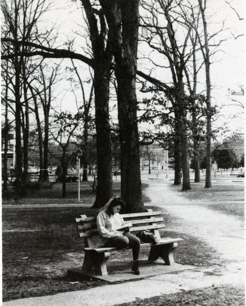 A female student, maybe around the early 1990s, sits on a bench with trees behind her. 