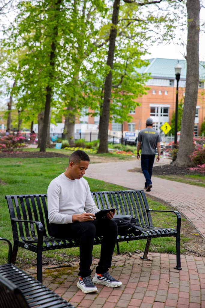 A male student sits on a bench with a tablet in his hands. 