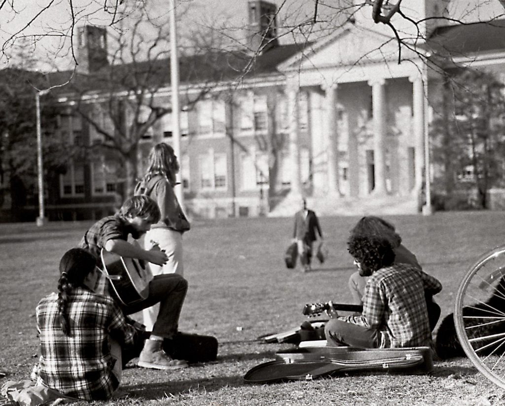 Five students sit on Bunce Green playing guitar and hanging out as a professor in the distance walks toward them. 