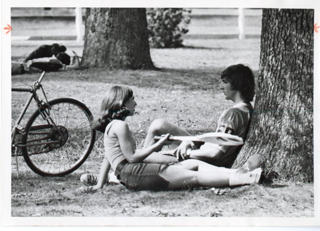 A female and male student, perhaps in the mid-1970s, relax together on the lawn, with her holding a tennis racquet and a bicycle to the side. 