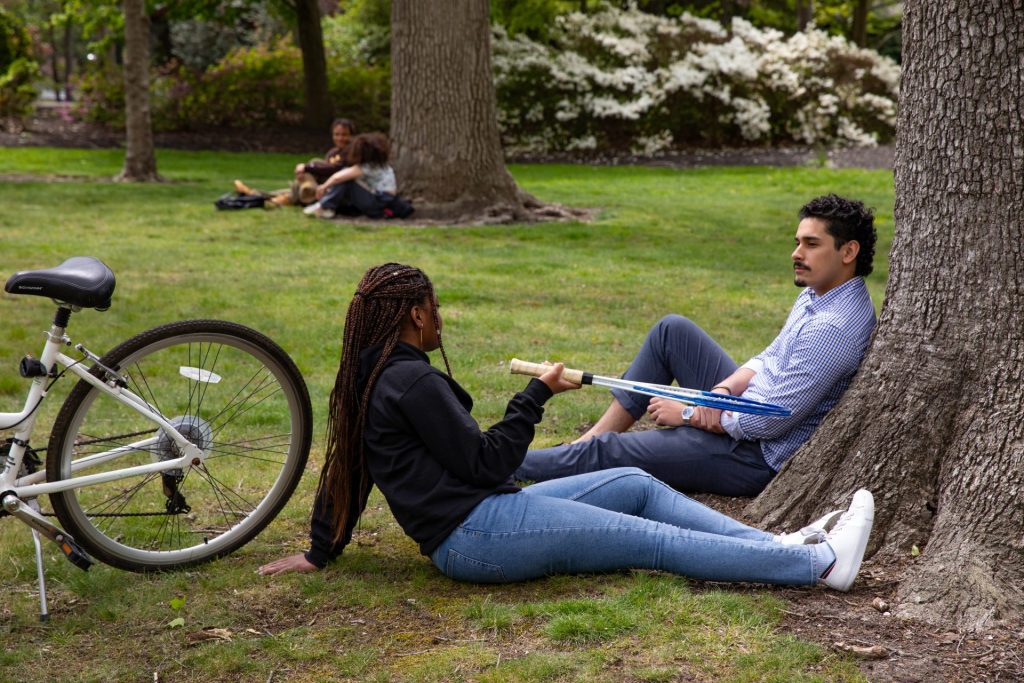 A female and male student, in 2023, relax together on the lawn, with her holding a tennis racquet and a bicycle to the side. 