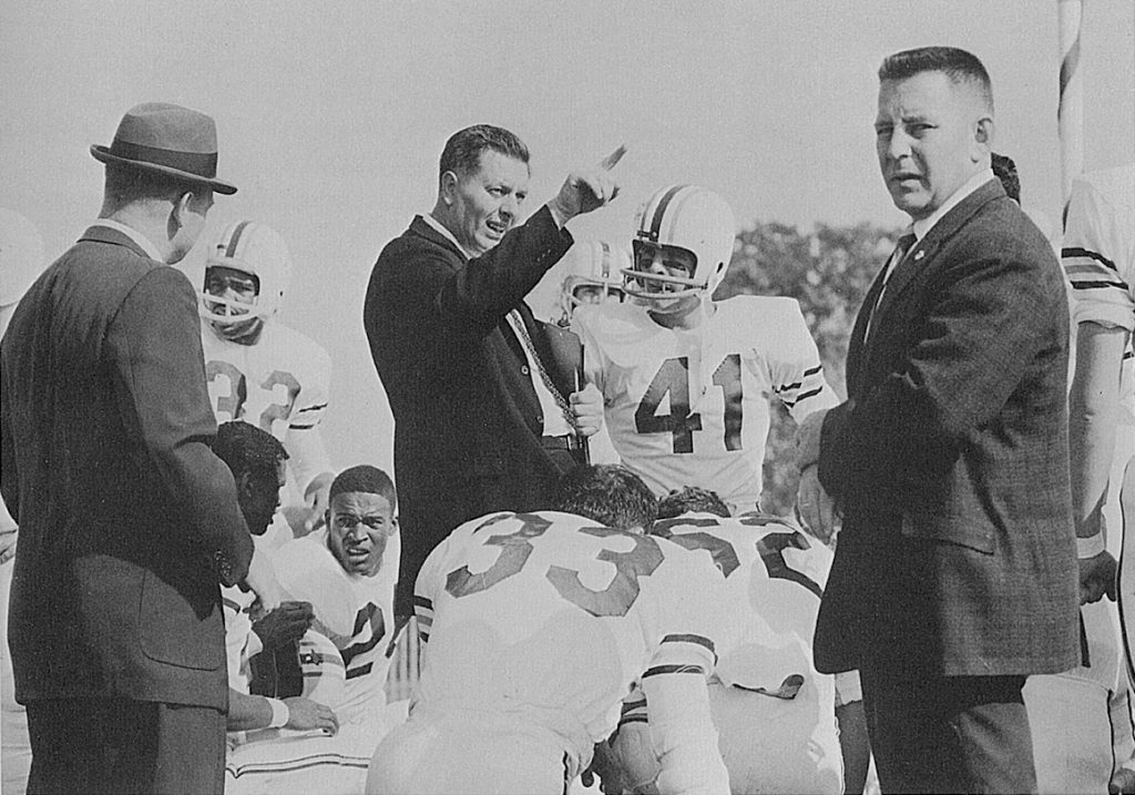 Football players and football coaches in the 1960s group together, with the coach pointing. 