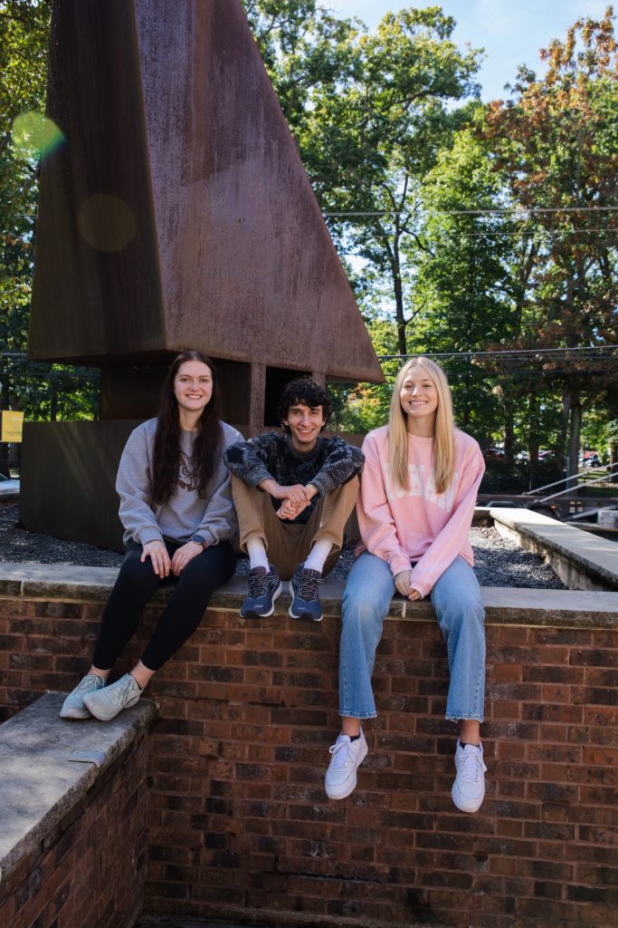 Three students in 2023 sit outside the Student Center in front of a sculpture. 